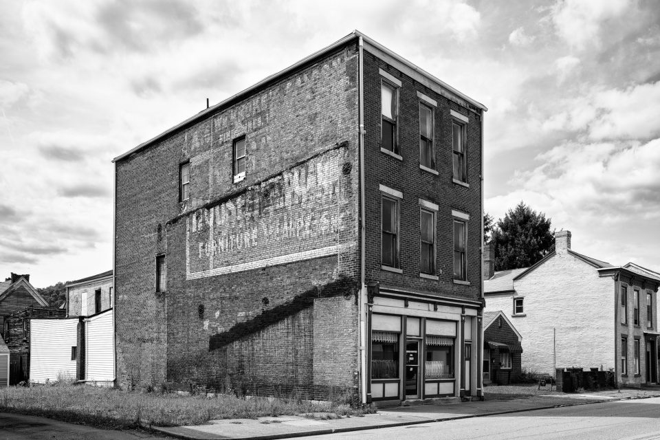 Black and white photograph of a three story brick building with layers of fading vintage wall ads on its side, seen in South Wheeling, West Virginia. Built in 1867, this the Stubenrauch Grocery with Stubenrauch Hall on the third floor. Click to buy a fine art print.
