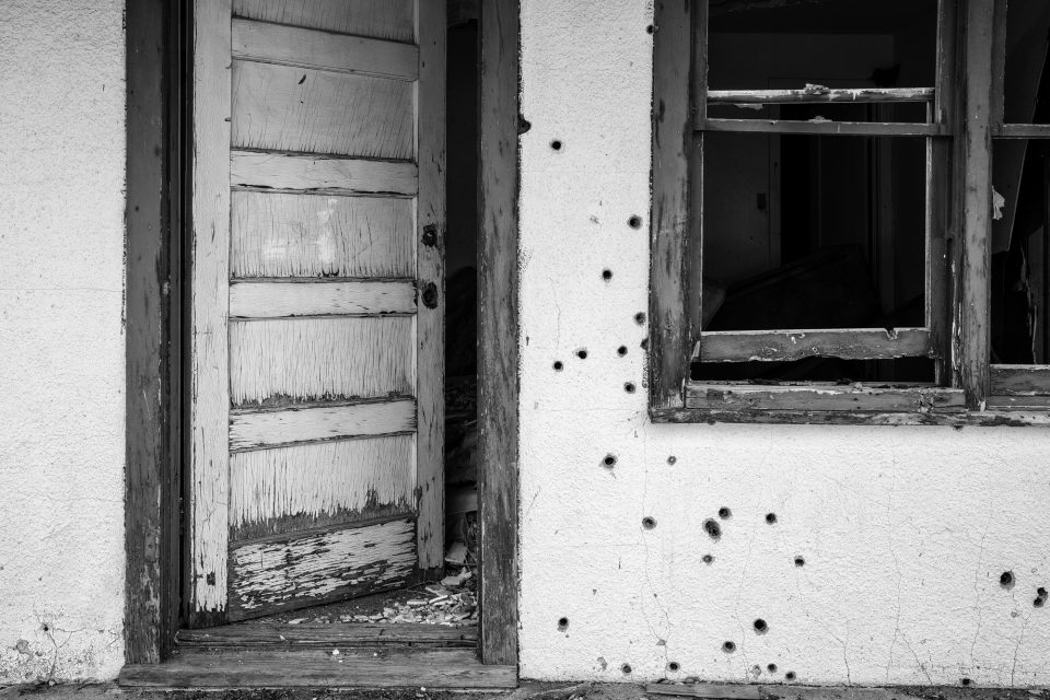 Black and white photograph of a stucco motel wall with bullet holes, by Keith Dotson.