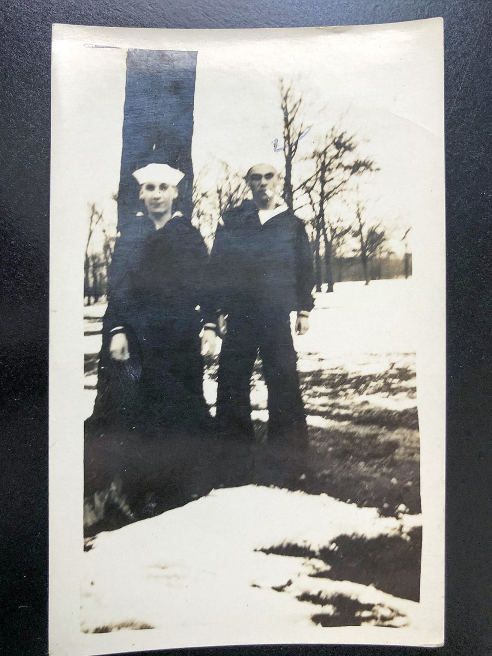 This undated old snapshot shows two young sailors in their uniforms. The snow and sky form a strong contrast with the black trees that their dark clothing blends into.