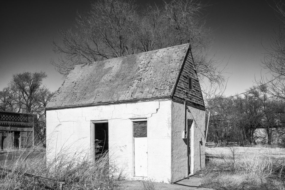 Black and white photograph of an abandoned building on the corner of Grand Avenue and Broadway in Texola. I think this would have been a corner gas station. The general merchandise store can be seen in the background.