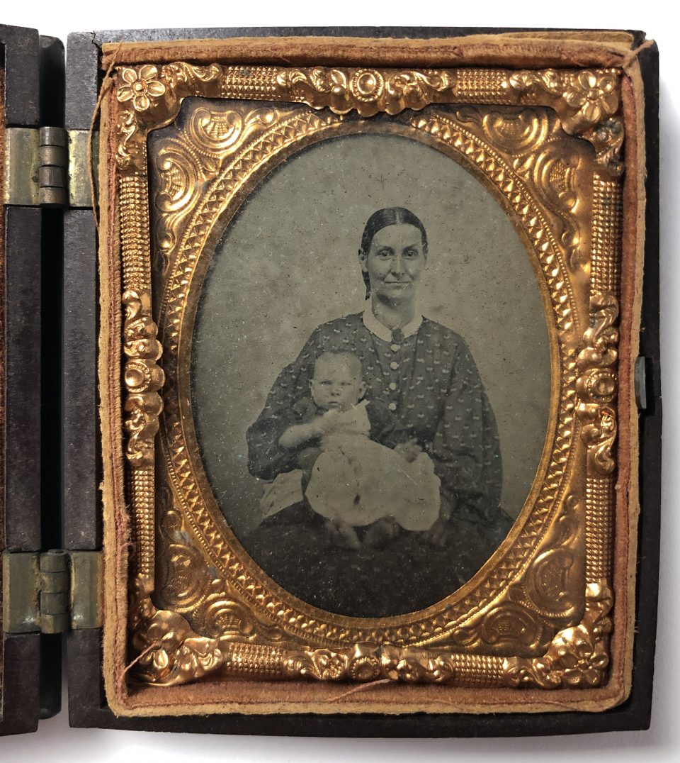 1860s tintype portrait of a smiling mother holding her baby on her lap. The shiny metal foil mats are made from stamped brass not much thicker than tin foil.