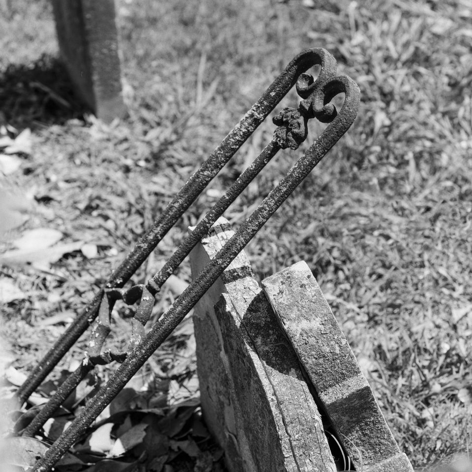 Piece of fallen iron fence leaning against old tombstones. Black and white photograph by Keith Dotson, shot with Hasselblad 500 C/M, with Cinestill Double X BwXX film.