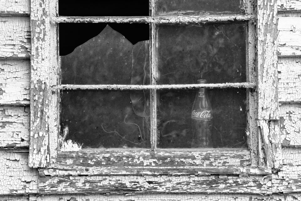 Black and white photograph of a side window of the old C.H. Pender Store, which shows broken glass and an empty Coca-Cola bottle 