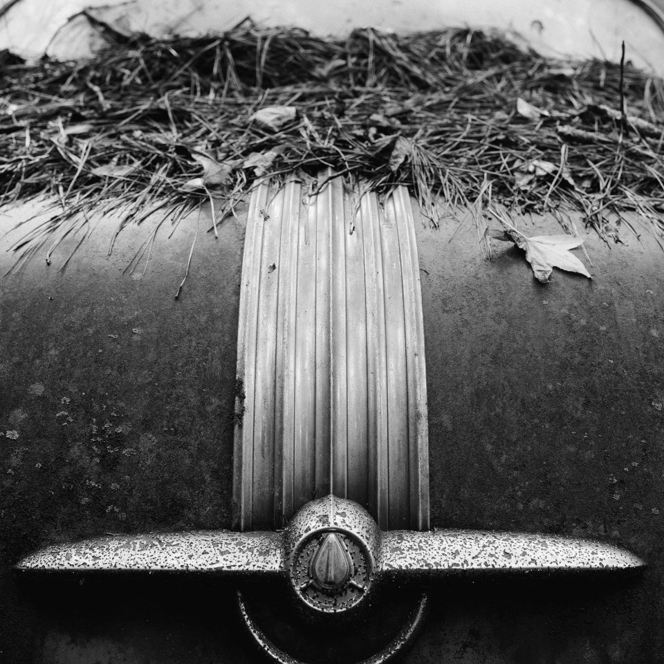 Ornament on Back of a Classic Car in a Junkyard - Black and White Photograph (Square Format) by Keith Dotson. Buy a fine art print.