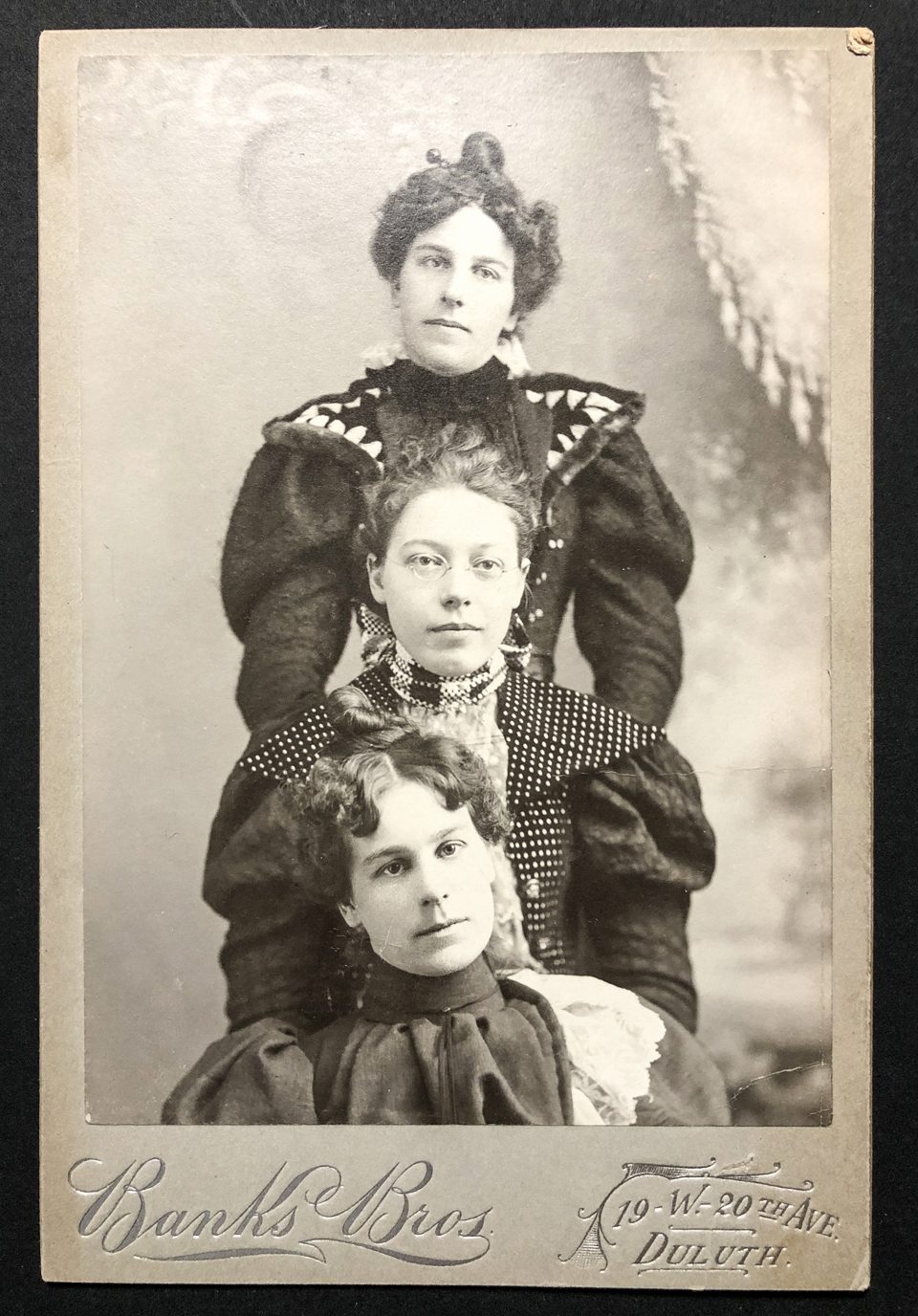 Cabinet card portrait of three young women in an unusual vertical lineup, made in the Duluth studio of the Banks Brothers. The studio name and address were stamped onto the front of the card with silver foil. The back of the card is blank. The photograph appears to be a gelatin silver print rather than an albumen print that was typically seen on cabinet cards.