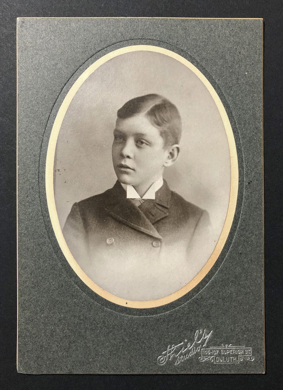 A head and shoulders portrait of a boy in a buttoned-up overcoat with an oval window mat by Theil’s Studio branding. The studio address was 105-107 Superior Street in Duluth, which places the date of this photo between 1891 and 1901.
