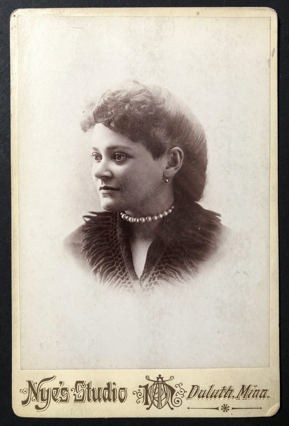 Head and shoulders portrait with a vignette effect of a young woman with a pearl necklace and fringed collar, made by the Nye's Studio of Duluth, Minnesota