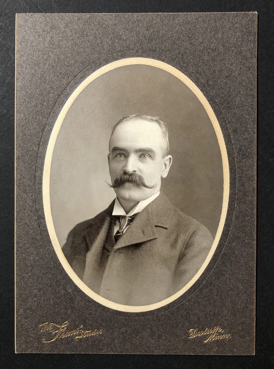 Black and white head and shoulders portrait of a man with a handlebar mustache wearing and wearing an overcoat, shot by The Fran's Studio in Duluth. The portrait is held in an oval photo mat, and the verso is blank.