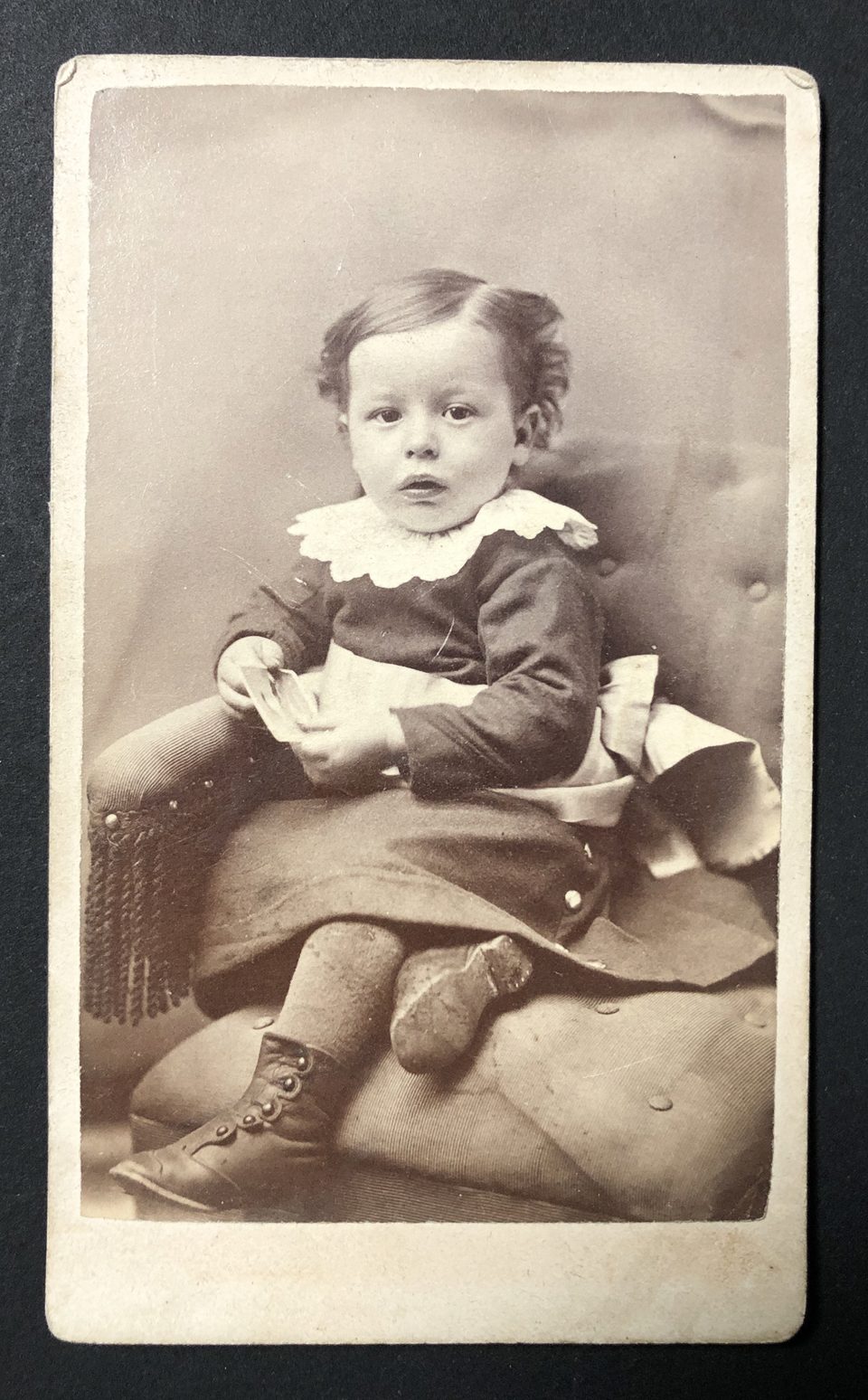 Portrait of a young girl wearing a long dress, thick leggings, and high, button-up boots. She's holding a photograph in her tiny hands. Notice the nails in the heel of her right boot.