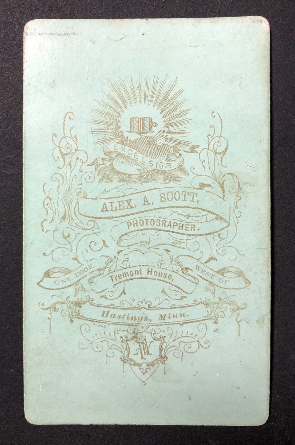 The back of this carte de visite is printed with gold ink on a robin's egg blue paper surface. It says "Excelsior. Alex A. Scott, photographer, One door west of Tremont House, Hastings, Minn."