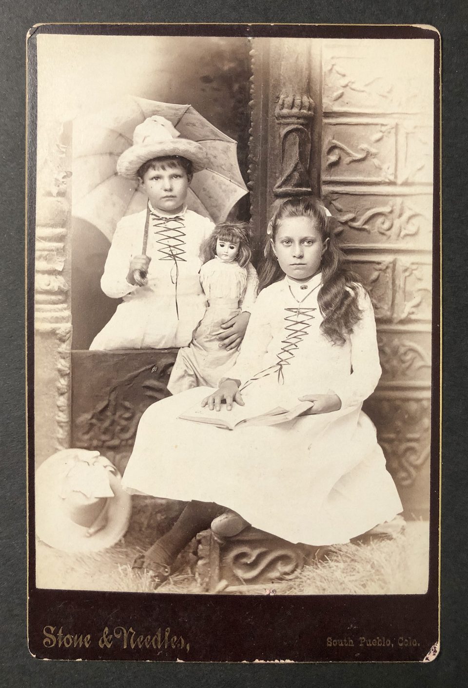 Cabinet card portrait of two young sisters wearing matching dresses, photographed by the studio of Stone & Needles in South Pueblo, Colorado. A close look shows that the younger girl moved slightly during the exposure.