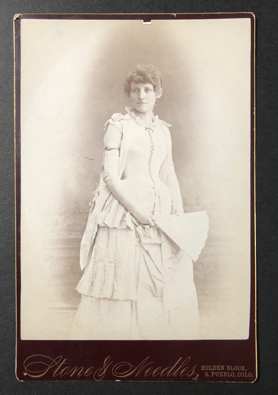 Cabinet card portrait of a young lady in a fancy white dress with long gloves and holding a fan, photographed by the studio of Stone & Needles in South Pueblo,