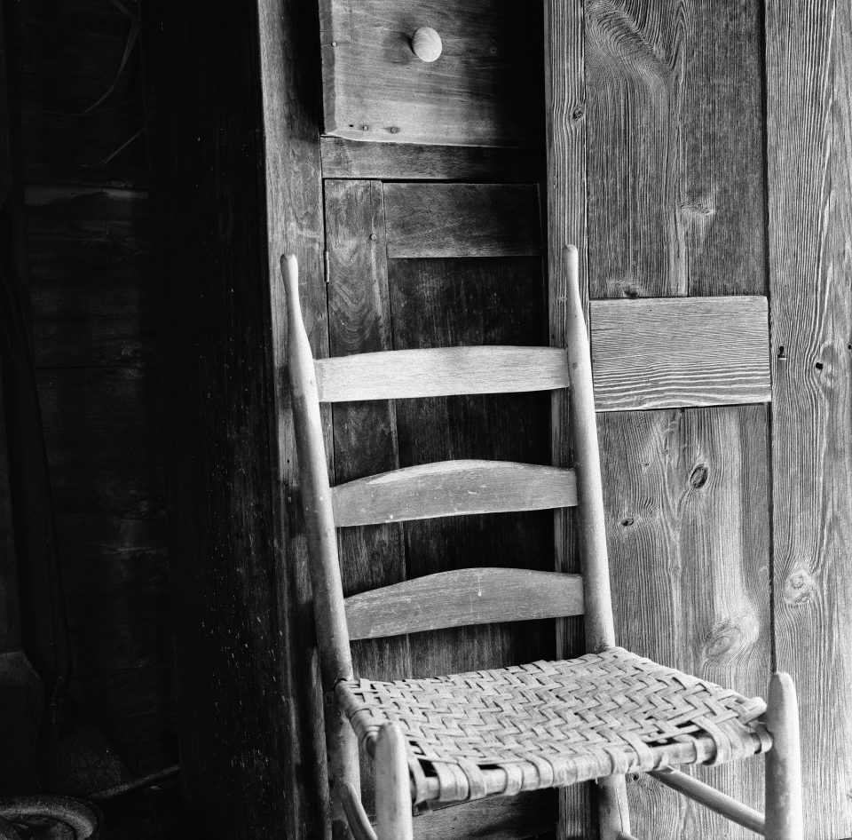 Black and white photograph of an old wooden chair inside a cabin at Museum of Appalachia in Tennessee, shot on Fomapan 100 film with Hasselblad 500 C/M camera by Keith Dotson.