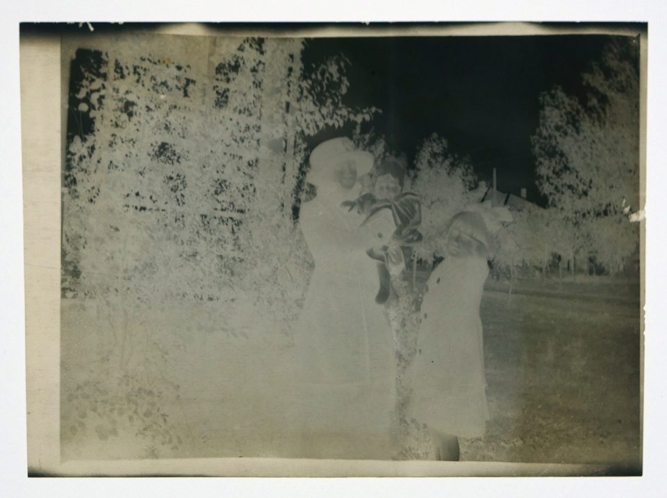 Antique film negative shows a woman with a baby and young girl in winter coats standing by a trellis.