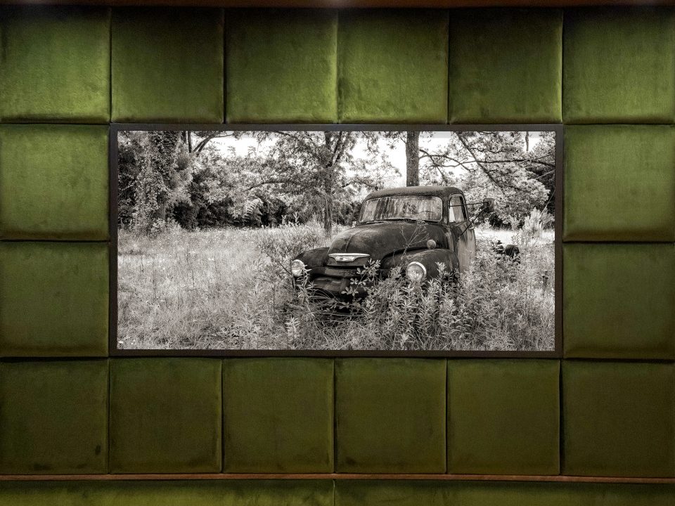 Large landscape photograph featuring an abandoned old pickup truck, rusting amidst tall grass and weeds. Photograph by Keith Dotson