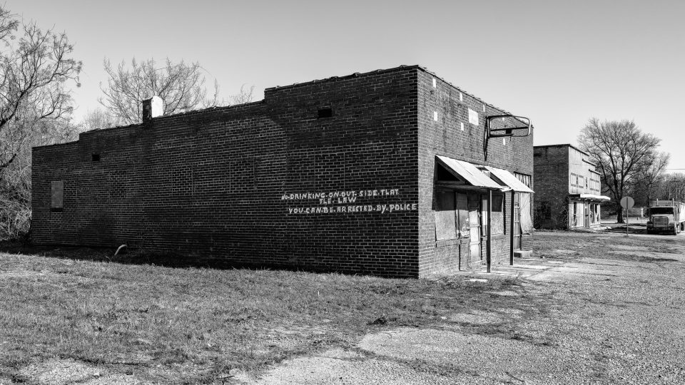Keith Dotson's black and white photograph of an abandoned building in Brinkley Arkansas, with a hand-painted sign that says "No Drinking on out side. That the Law. You Can Be Arrested by Police."