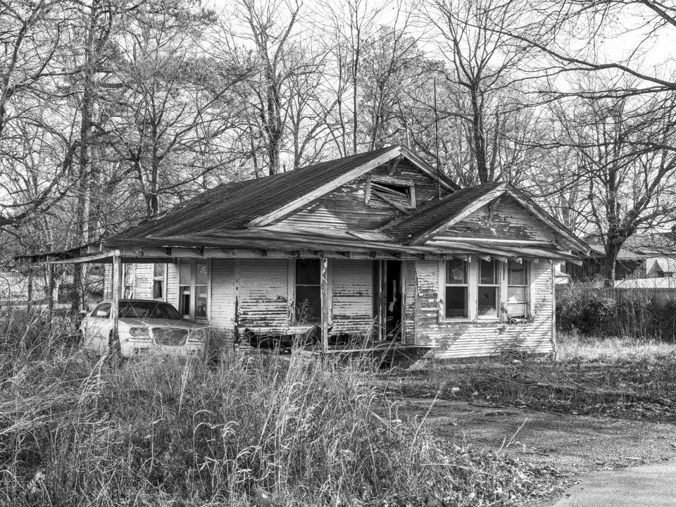 This old house is a shambles, standing wide-open to the elements, but there's a nice car under the car port. Black and white photograph by Keith Dotson.