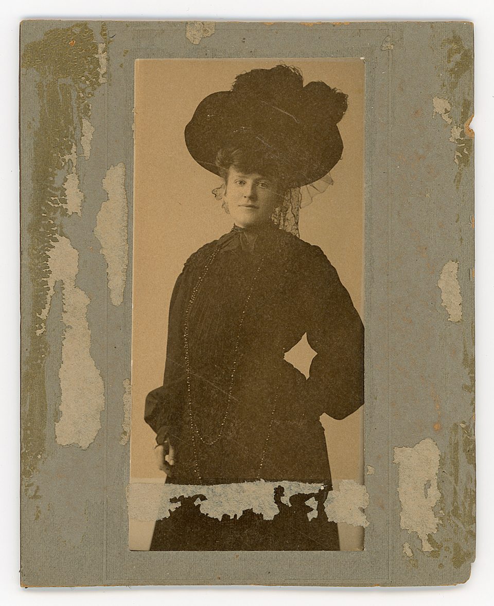 Small cabinet card portrait of a young woman stylishly dressed in Edwardian-style popular fashion, including a big hat with feathers, puffy sleeves, and very long beaded necklaces. Made in the early 1900s by Japanese-American photographer Tokujiro (Henry) Motoyoshi at his studio located at Stockton and O'Farrell streets in San Francisco.