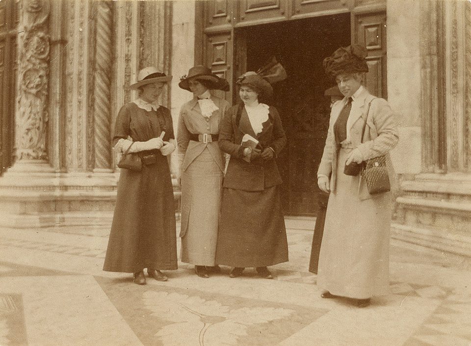 "In front of the Duomo, Siena." 4-inches wide by 3-inches tall. Sepia toned gelatin silver photograph, circa 1910. The photograph shows five well-dressed tourists in "Merry Widow" hats and tailored Edwardian walking suits. Photographer not identified.