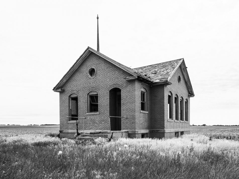 Black and white photograph of a red brick one-room schoolhouse standing abandoned on the Kansas prairie.