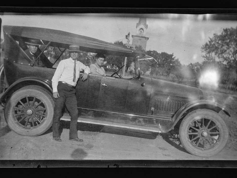 Photograph of two men wearing straw boater hats and a woman seated inside an early 1900s automobile. In the background is visible of what appears to be a church steeple.