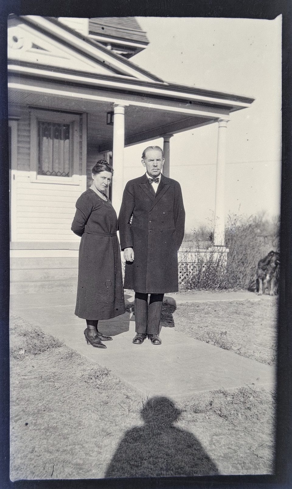 Portrait of a middle-aged man and woman in their dress clothes standing outside the farmhouse, with the photographer's shadow visible in the frame. Their facial expressions appear to be stern.