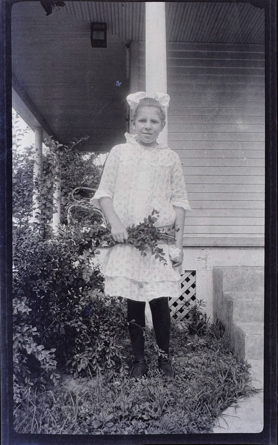 Portrait of a young girl holding flowers outside her house. AI dated this photograph to about 1900.