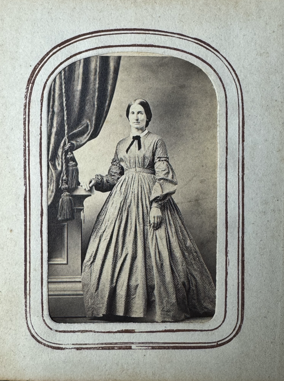 Beautiful, full length portrait of a woman in a spotted dress. We estimate this carte de visite portrait to be from 1860 - 1865, based on her full, bell-shaped skirt, narrow waist, and large pagoda-style sleeves. Her center-parted, smooth hair gathered toward the back is also compatible with the Civil War era. The elaborate curtain, pedestal, and painted studio backdrop are also typical of CdV portraiture of the 1860s.