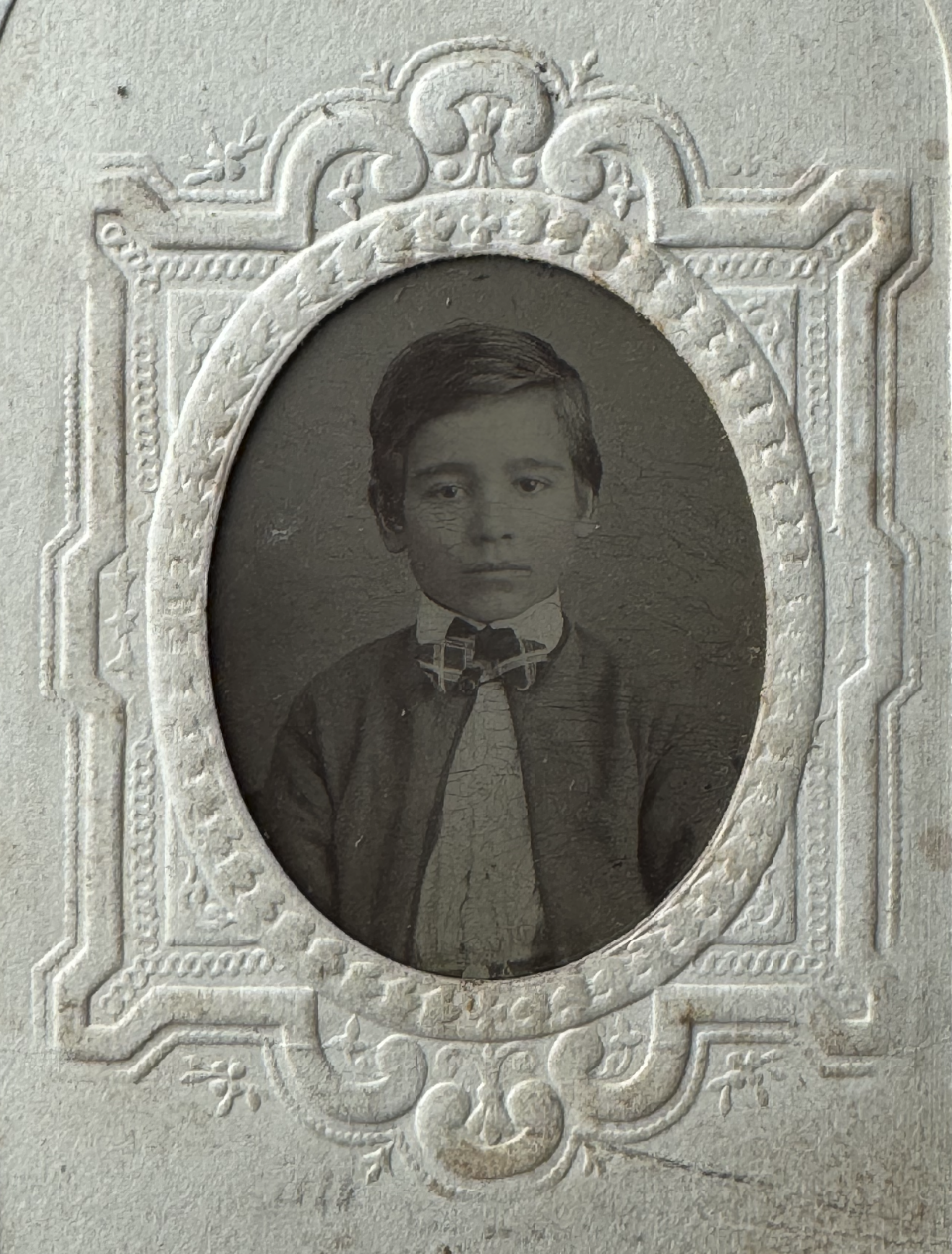 Tintype portrait of a boy with a clean cut hairstyle wearing a plaid bowtie. Note the ornate embossing of the paper mat.